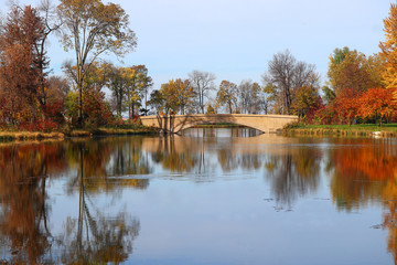 Fototapeta na wymiar Beautiful fall landscape with a bridge in the city park.Scenic view with colored trees around old style bridge in sunlight reflected in the lake Mendota bay water. Tenney Park, Madison, Wisconsin, USA