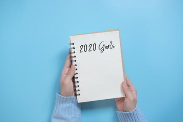 New Year 2020 goals list with notebook on blue background, flat lay style. Planning concept.