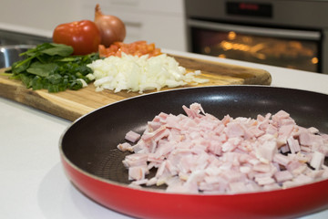 Home Kitchen With Bacon Cut Into Pieces In Pan And Veggies On A Chopping Board With Oven In Background