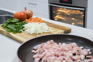 Home Kitchen With Bacon Cut Into Pieces In Pan And Veggies On A Chopping Board With Oven In Background