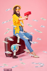 Smiling woman traveler sitting on luggage holding money gun model in holiday on pink backgrounds, relaxation concept, travel concept