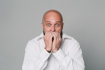 Handsome serious middle-aged man in a white shirt looks at the camera in surprise and holds his hands near his face on a gray background