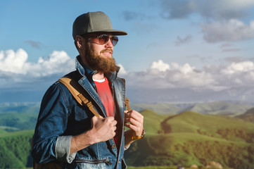Portrait of a stylish smile young man with a long beard in a jeans suit, sunglasses and a cap with a backpack stands on a background of mountain valley in nature.
