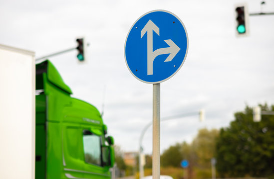 A blue and white round sign in Germany shows the directions straight ahead and right. Traffic lights show green and a truck drives by.