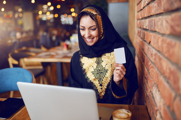 Beautiful smiling muslim woman sitting in cafe, holding credit card and looking at laptop. On wooden desk are coffee and laptop.