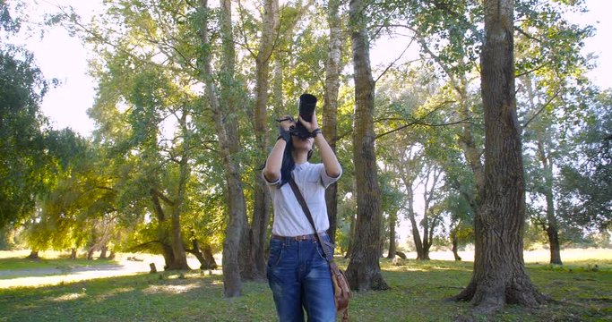 Steady around slide camera shot attractive female  photographer  taking  pictures of nature with big dslr camera in a forest belt of large poplars in Europe on sunny day.  4k slow motion 50 FPS