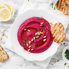 Baked Beet Hummus with toasted bread in a white ceramic plate on a white wooden background. Top view
