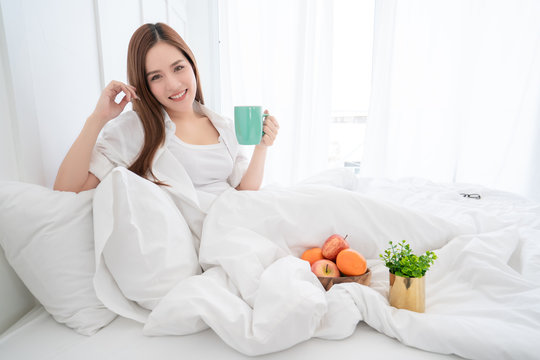 beauty asian female white dress long hair hand hold hot drink morning time  on white bed with clam and cheerful white bedroom interior background