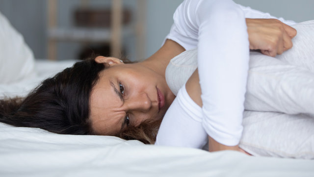 Depressed young mixed race woman lying on bed in bedroom, embracing knees.