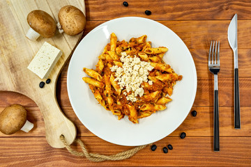 Top view of fresh cooked vegetarian pasta noodles with red sauce, tofu on white plate with fresh mushrooms, fork, knife and black soybeans around, on wooden table.