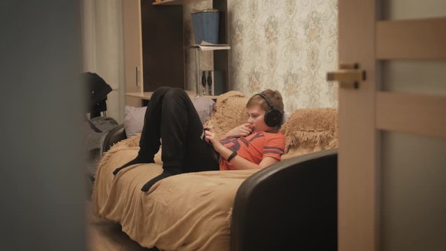Boy watching media content using headphones and mobile phone app lying on the couch at home. Close Up of boy hands playing video games on cell phone in the evening.