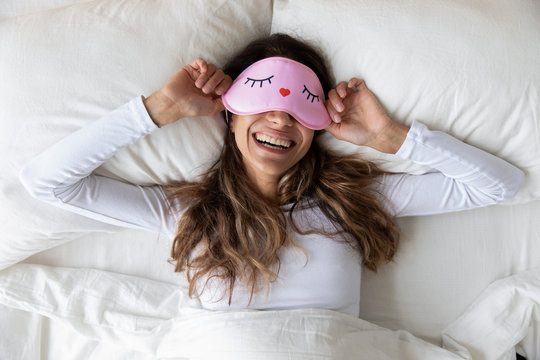 Happy millennial woman lying in bed with sleeping mask.