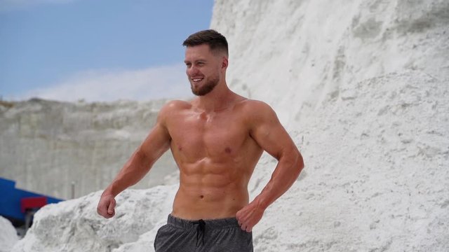 Muscular bodybuilder smiling outdoors. Strong athlete shows his trained body while standing on the white hill.