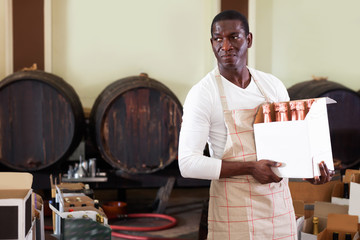 Positive afro seller working in wine store, holding box full of bottles with wine