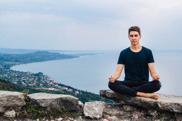 Naklejka na ściany i meble Young handsome man in a meditative position sits against the background of the sea, mountains and city. Concept of freedom relaxation. Place for text or advertising