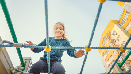 A charming little girl in casual clothes sits on the grid and looks at camera on playground in the...