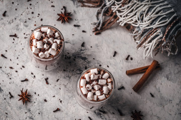 Two glasses of hot cocoa with marshmallows and chocolate chips on a background of scattered cloves, chocolate, star anise and cinnamon, with a blanket in the background