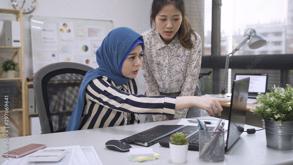 Wall mural Irritated young asian muslim woman manager working angry with chinese fresh graduate. muslim female leader point finger at laptop screen asking strict japanese intern of mistake in project in office