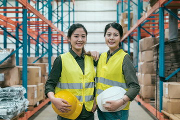 Cheerful woman coworkers of warehouse smiling and looking at camera. confident lady colleagues in stockroom posing with carrying hard hat. best team work partner in storehouse lifestyle concept.