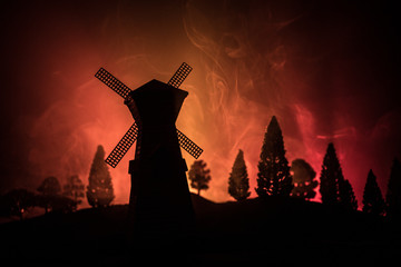 Windmill silhouette standing on hill against the night sky. Night decor with old windmill on hill with horror toned foggy background with light.