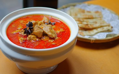 Red chicken curry with anises in ceramic bowl and fried flatbread in round bamboo plate on wooden table