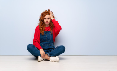 Redhead woman with overalls sitting on the floor with an expression of frustration and not understanding