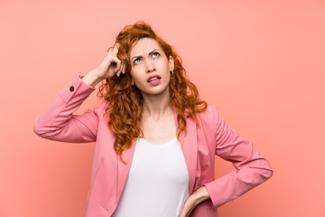 Redhead woman in suit over isolated pink wall having doubts and with confuse face expression