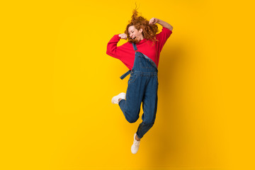 Redhead woman with overalls jumping over isolated yellow wall