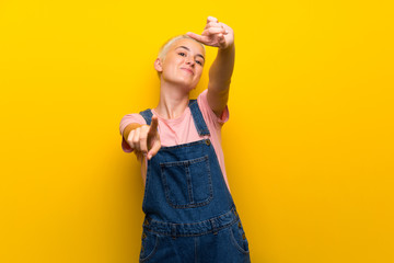 Teenager girl with overalls on yellow background points finger at you while smiling