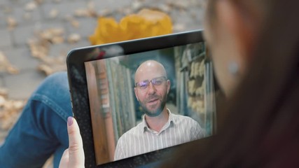 Caucasian woman video calling with bald bearded male friend using tablet on the bench in park. Female shown from behind speaking with man through video chat from autumn park.
