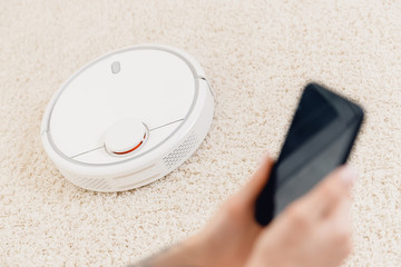 Young girl using automatic smart robot vacuum cleaner working on carpet. Controlling device with phone
