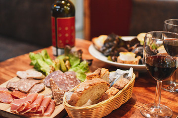 Spanish tapas meal with basket of bread, plate of cold cut meats, chèvre cheese salad and glasses of red wine. All on wooden table -Image - obrazy, fototapety, plakaty