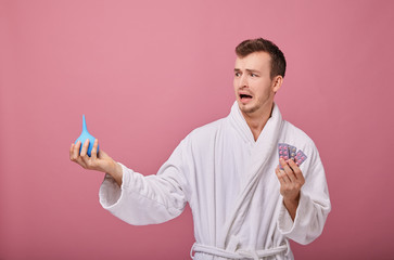 A man with poor health stands on pink background and holds an enema in his right hand and in the left pills. The guy looks fearfully at the medications. Recovery, treatment, procedures, poor health.