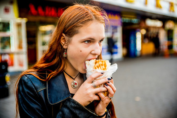 portrait of young teenager redhead girl with long hair eating chicken shawarma on street