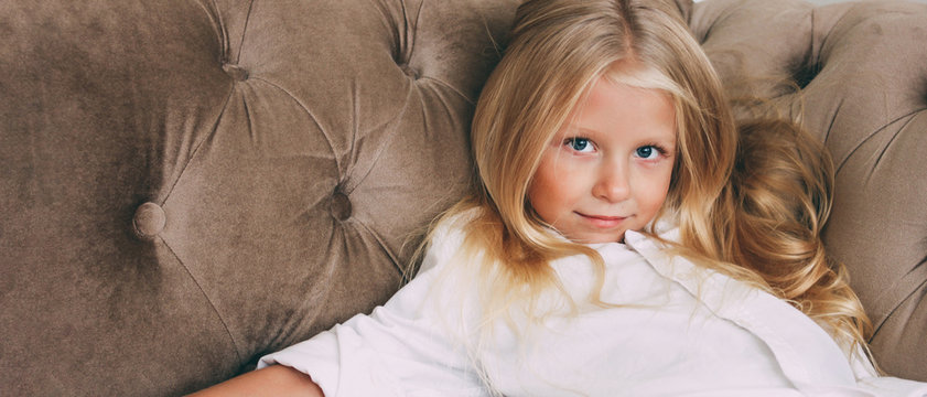 Beauty fashion portrait of smiling little tween girl with fair long hair in white shirt on beige sofa background banner, children's modeling
