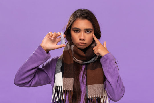 Pretty african-american girl in a sweater and cosy checked scarf using nasal spray receive treatment for running nose standing isolated over purple background.