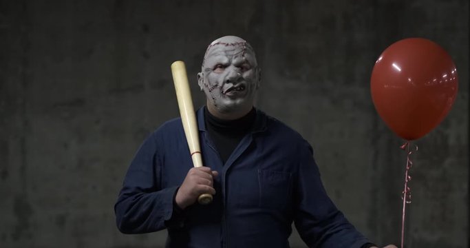 Extremely dangerous man in mask of movie maniac imposingly goes to camera, psycho in parking lot, Halloween holiday, creepy costume, October walk, red ball and bat in his hand, alien from hell