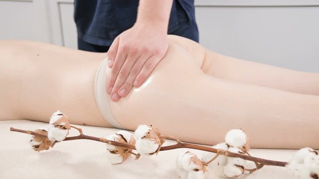 Close-up of a male physiotherapist rubbing massage oil on the thigh and buttocks of a young woman before massage treatments in a professional spa salon. Concept of relaxation and body care in 4k