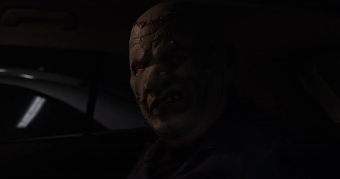 Shocking monster is sitting in backseat of car in dark parking lot, ambush, unpleasant surprise for driver, maniac waiting for victim, Halloween costume, creepy image, disgusting mask, half darkness