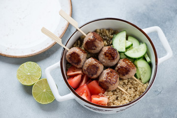 Serving pan with with meatball skewers, quinoa, bulgur and fresh vegetables, elevated view on a light-blue stone surface