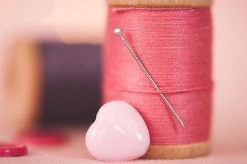 Pink cotton thread spool with heart shape button and needle closeup. Selective focus.