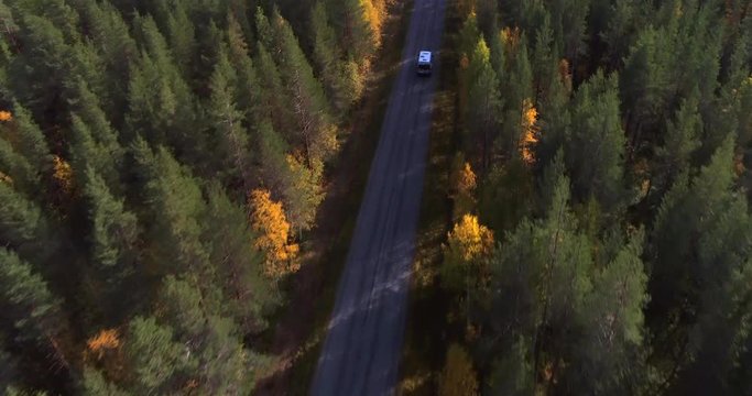 Camper on a forest road, Aerial, drone shot, overlooking a camper van, driving on a asphalt route, in middle of autumn colored trees, on a sunny, ruska day, in Pyha-Luosto national park, Finland