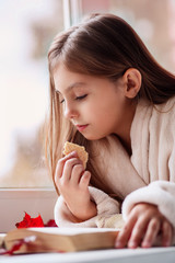 Cute little girl lying on the window, eating cookie and reading a book at home