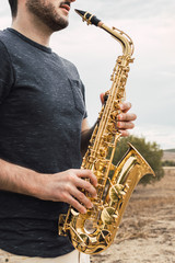 Obraz premium Young bearded man plays saxophone in a natural place during a cloudy day in the countryside. Playing musical instrument in the nature.