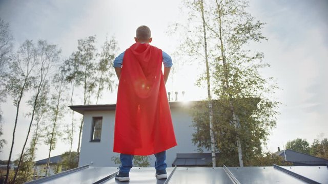 Boy is Playing a Role of a Super Hero. He's Standing on a Roof of a House with His Hands on His Waist. Young Man is Wearing a Bright Red Cape. He's Looking at the Sun.
