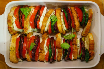 Flat lay top view of oven baked sliced vegetables (tomatoes, aubergines and courgettes) with mozzarella cheese and baguette slices dressed with basil.