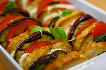 Flat lay top view of oven baked sliced vegetables (tomatoes, aubergines and courgettes) with mozzarella cheese and baguette slices dressed with basil.