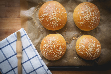 Top view of homemade sesame buns on a wooden chopping board with a striped white kitchen cloth and a pastry brush.