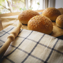 Top view of homemade sesame buns on a wooden chopping board with a striped white kitchen cloth and a pastry brush.