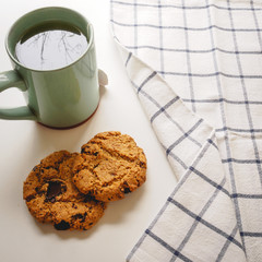 Chocolate chip cookies and a green mug of tea with a tea towel on a white table. Flat lay. Square format